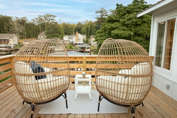 Two wicker chairs with cushions on a wooden deck, a small white table, and a view of houses and trees in the background.