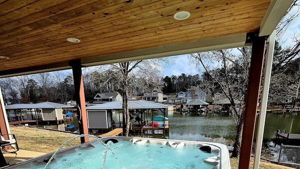 Un bain à remous sous une pergola en bois, avec vue sur un lac et des maisons en arrière-plan.
