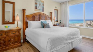 Ein Hotelzimmer mit einem großen Bett, einem Holzkopfstück, zwei Nachttischen mit Lampen, einer Kommode und einem großen Fenster mit Blick auf den Strand.