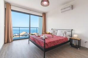 A bedroom with a large window, a bed with a red patterned bedspread, a bedside table with a lamp, and a view of the sea. - Casa Teixeira by Atlantic Holiday (Calheta)