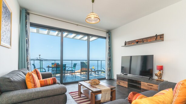 Un salon moderne avec un canapé, une table basse et un téléviseur. Il y a un balcon avec vue sur la mer.