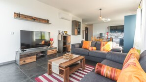 Un salon moderne avec un téléviseur à écran plat, une table basse en bois, un canapé gris agrémenté de coussins colorés, et une cuisine en arrière-plan.