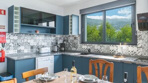 Une cuisine moderne avec des meubles bleus, une grande fenêtre donnant sur les montagnes, un pare-feu en carrelage et un coin repas avec une table dressée pour deux personnes.