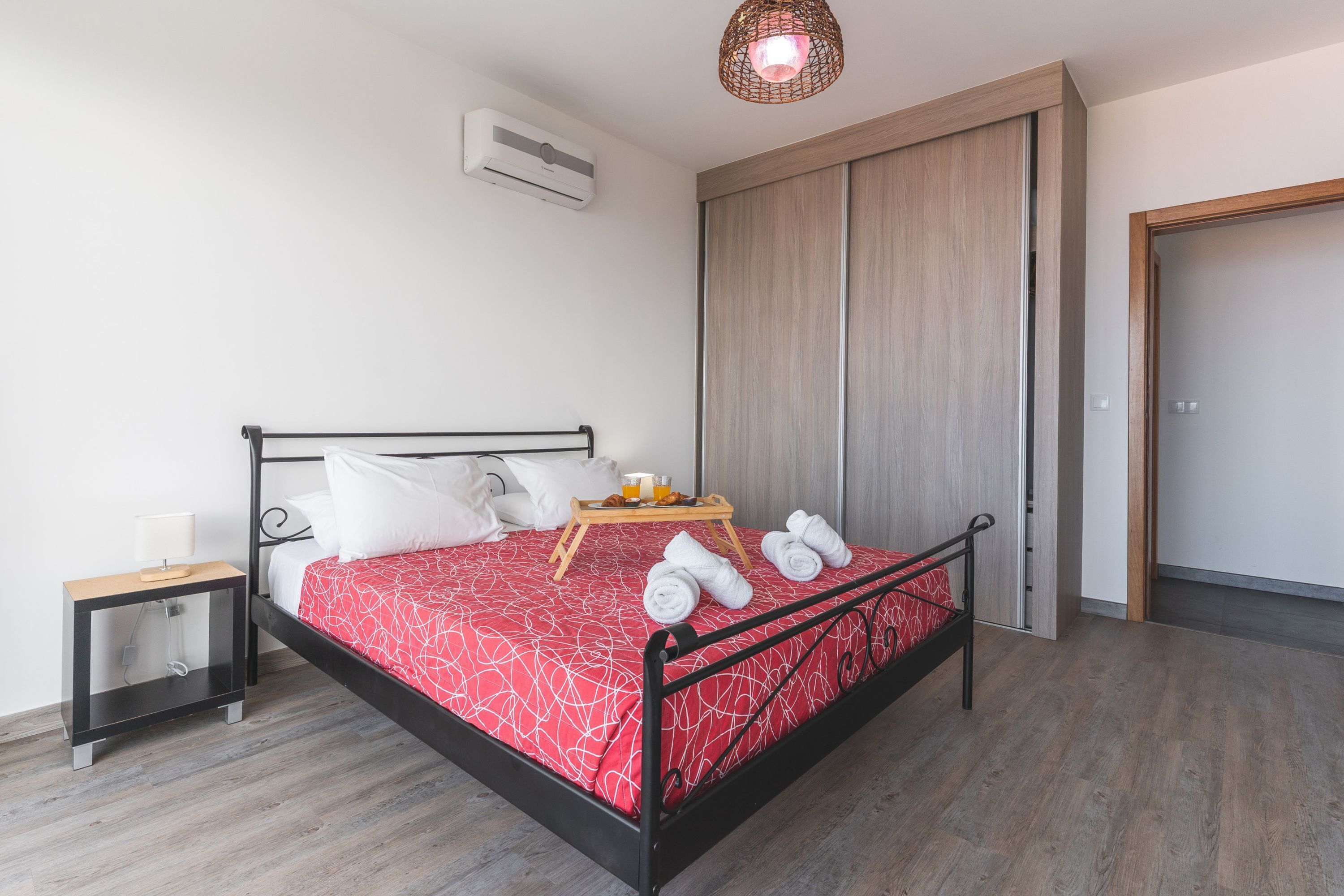 A neatly made bed with a red and white patterned bedspread, a tray with towels, and a bedside table with a lamp.