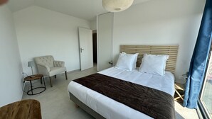 A modern bedroom with a large bed, a chair, a lamp, and a view of the outdoors. - Le Toit Penché (Tardinghen)