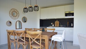A dining area with wooden chairs and a white table, a kitchen with white cabinets and a black countertop, and three hanging pendant lights. - Le Toit Penché (Tardinghen)