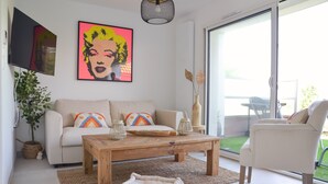 A living room with a sofa, coffee table, and a chair. There is a TV mounted on the wall and a window with a view of the outdoors. - Le Toit Penché (Tardinghen)