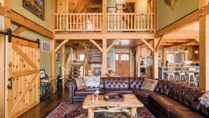 A spacious living area with a brown leather sofa, a wooden coffee table, and a patterned rug. A staircase leads to an upper level with a balcony. - Spacious Finger Lakes Waterfront Lodge (Road Burdett)