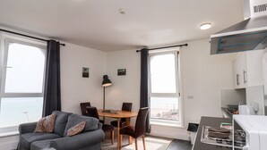 Ein gemütlicher Wohnraum mit einer Couch, Esstisch, Stühlen, einer Lampe, einer Küchenzeile und großen Fenstern mit Blick.