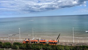 Blick auf die Küste mit einem Sandstrand, einer Promenade mit orangefarbenen Sitzgelegenheiten und blauem Himmel.