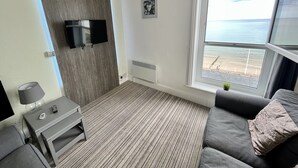 Ein modernes Zimmer mit großem Fenster und Blick auf den Strand, einem an einer Holzvertäfelung befestigten Flachbildfernseher, einem grauen Sofa mit gemustertem Kissen und einem kleinen Tisch mit Lampe und Fernbedienungen.