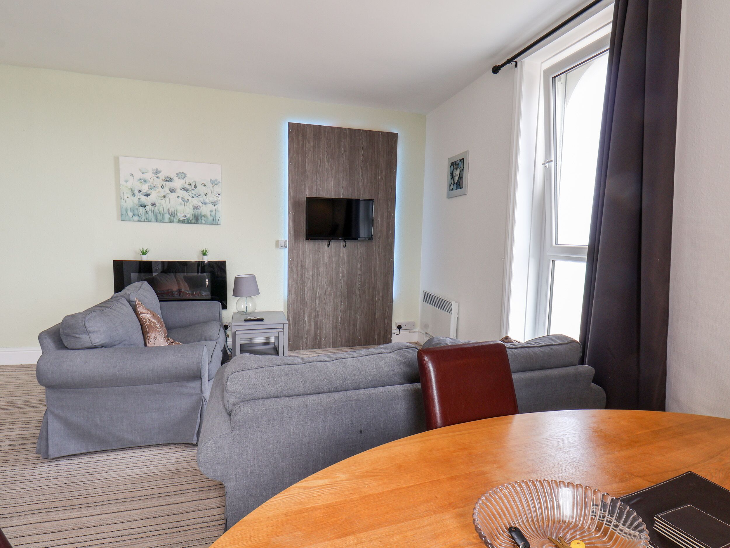 Ein Wohnzimmer mit grauem Sofa, einem Holzesstisch und einem an der Wand befestigten Fernseher.