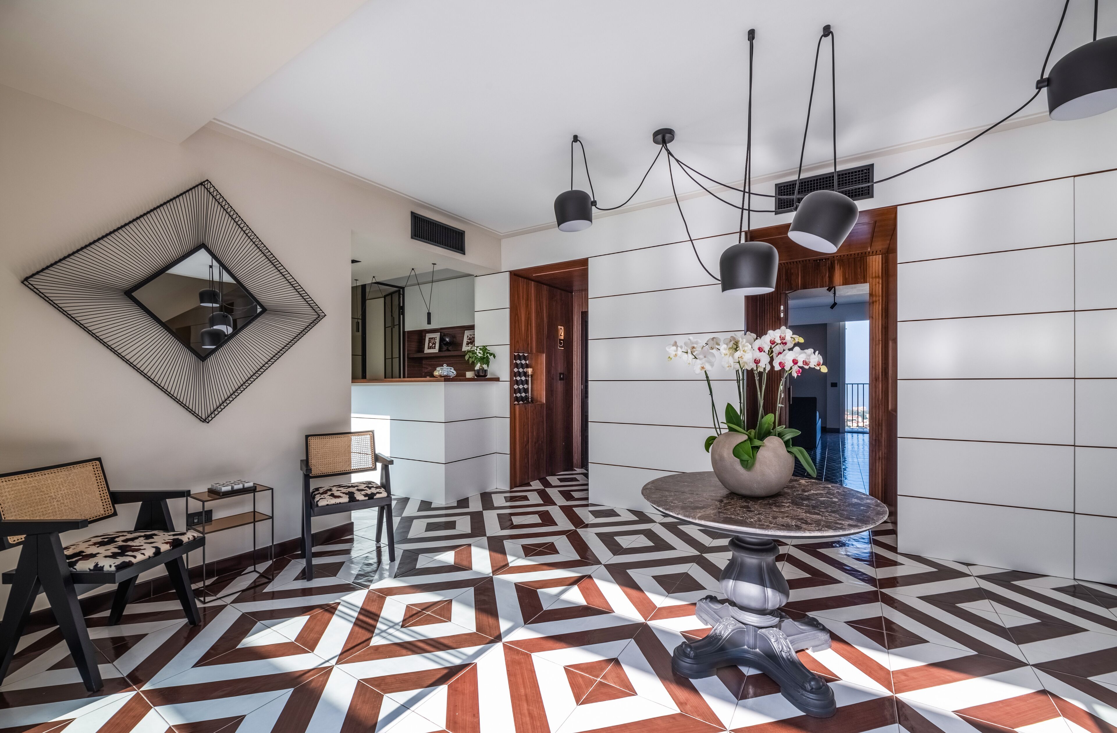 Un vestíbulo moderno con suelo de patrón geométrico, una mesa redonda con un jarrón de flores y un espejo grande en la pared.