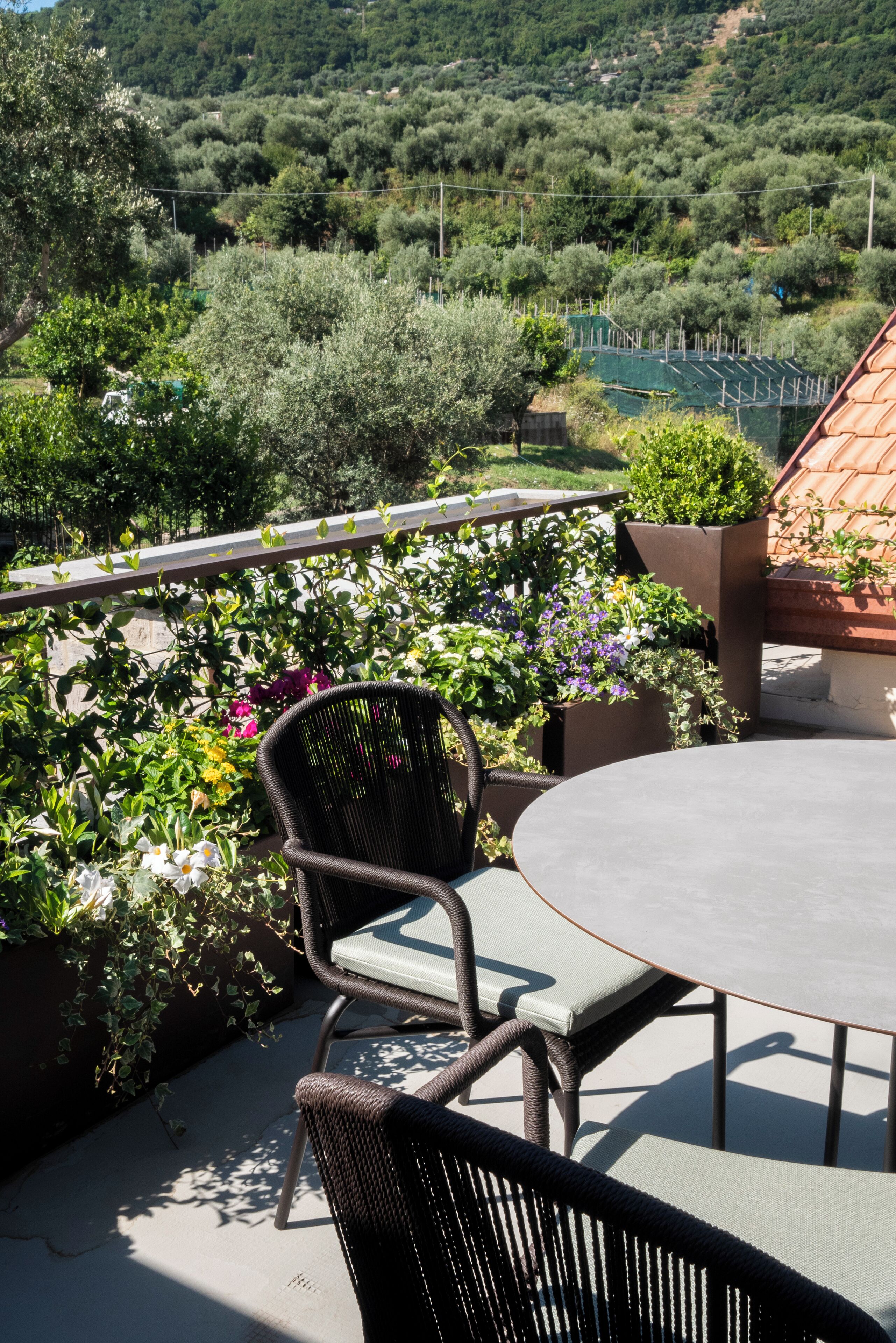 A round table with two wicker chairs, surrounded by lush greenery and a view of a vineyard.