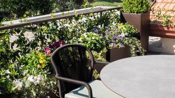 A round table with two wicker chairs, surrounded by lush greenery and a view of a vineyard.