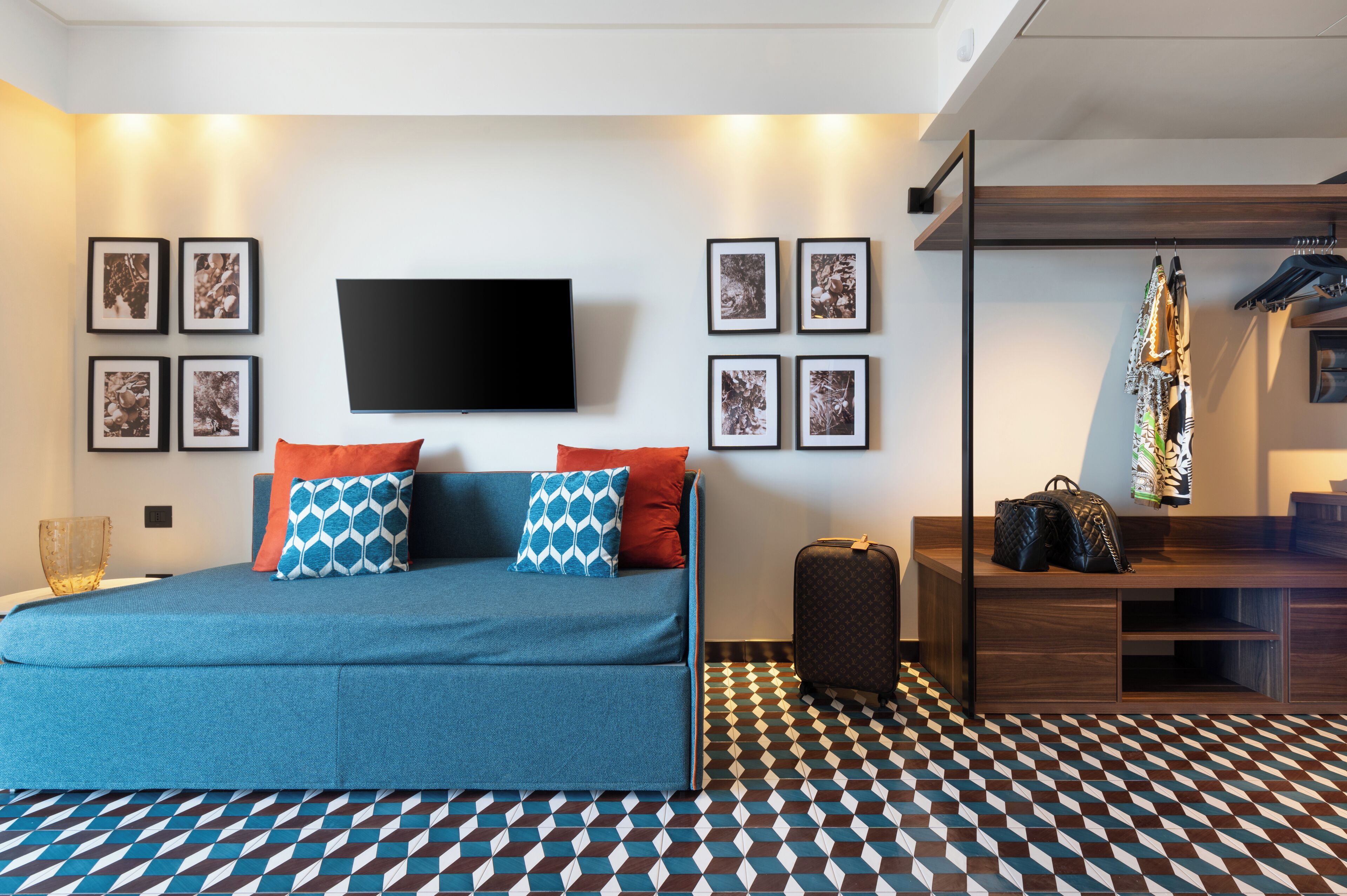 A modern bedroom with a sofa bed, a flat-screen TV, framed pictures, a patterned floor, and a wooden bench with bags.