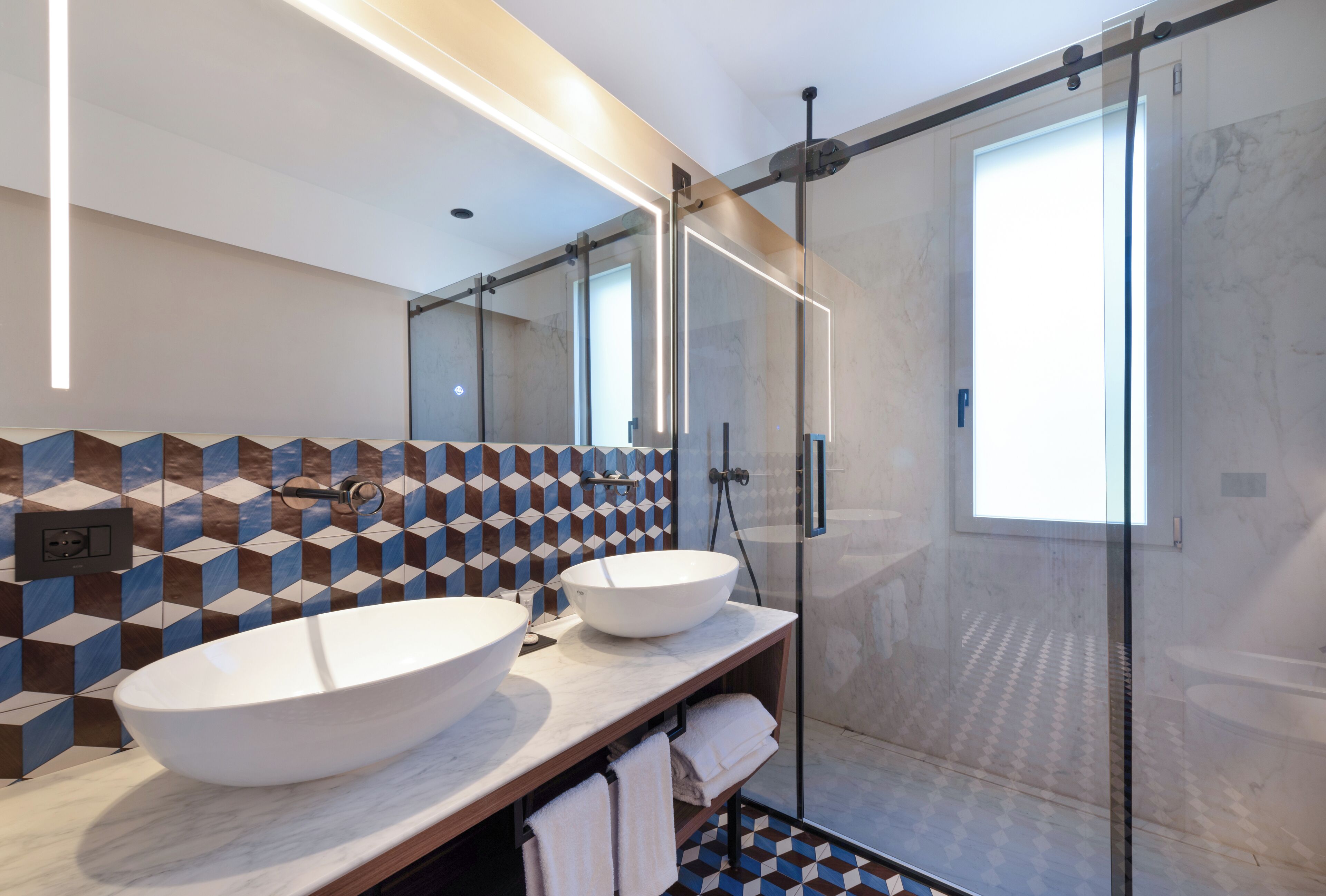 A modern bathroom with a large mirror, two sinks, a glass shower enclosure, and patterned tile walls.