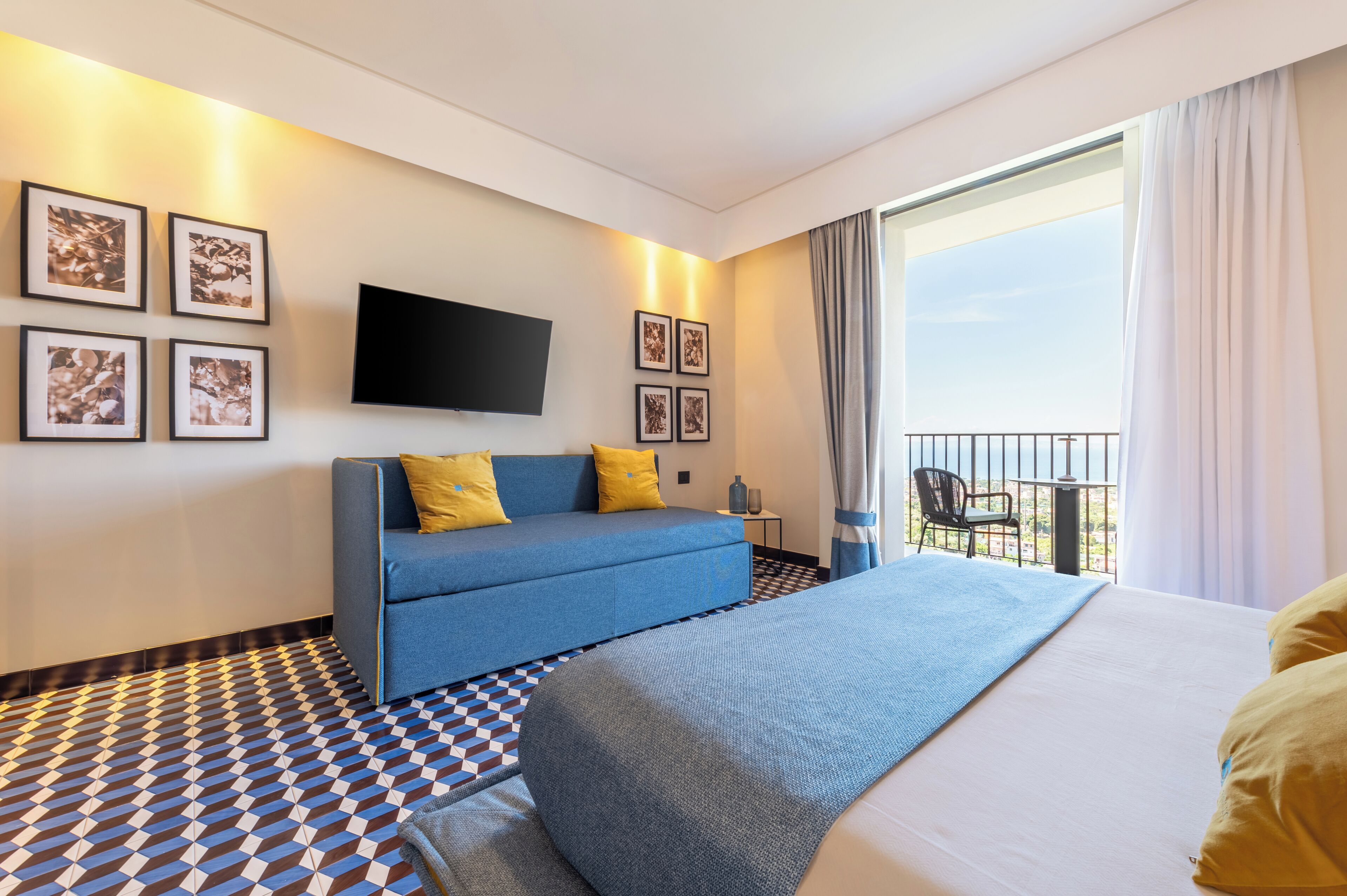 Ein Hotelzimmer mit einem großen Bett, einem blauen Sofa, einem Flachbildfernseher und einem Balkon mit einem Stuhl.