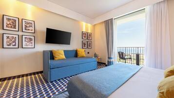 Ein Hotelzimmer mit einem großen Bett, einem blauen Sofa, einem Flachbildfernseher und einem Balkon mit einem Stuhl.