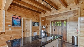 Interior de una cabaña de madera con cocina, que incluye encimera de granito, pileta de acero inoxidable y armarios de madera.