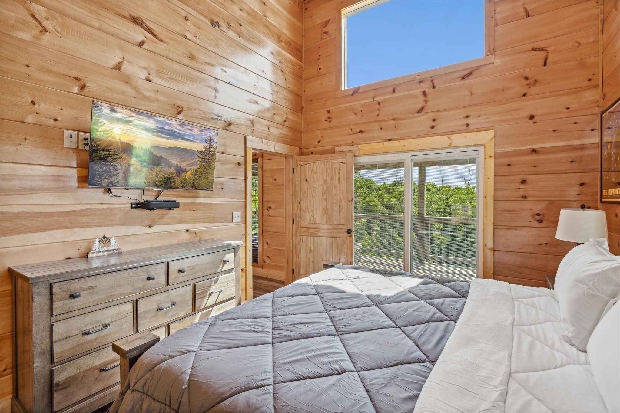 A wooden cabin bedroom with a bed, dresser, TV, and a window with a view of greenery.
