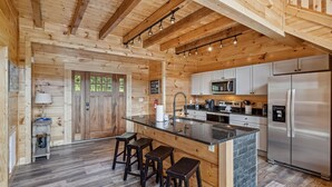 Interior de una cabaña de madera con una zona de cocina que incluye un refrigerador de acero inoxidable, un microondas y un lavamanos. Hay tres taburetes en la barra y una puerta de madera con ventana.