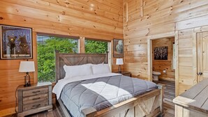 Un dormitorio tipo cabaña de madera con cama, mesitas de noche, ventana con vistas y puerta que da a otra habitación.