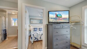 Un dormitorio con cama, cómoda, televisor mostrando 'Seaside Vacations & Sales' y ventana con persianas.