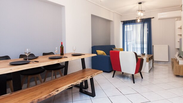 A modern living room with a dining area, featuring a wooden dining table, black chairs, a blue sofa, and a red and white chair. - Home Away from Home 2 Bd Apt in Zografou (Zografos)