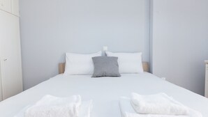 A neatly made bed with white bedding and a grey pillow, two folded white towels at the foot of the bed, and a plain light-colored wall. - Home Away from Home 2 Bd Apt in Zografou (Zografos)