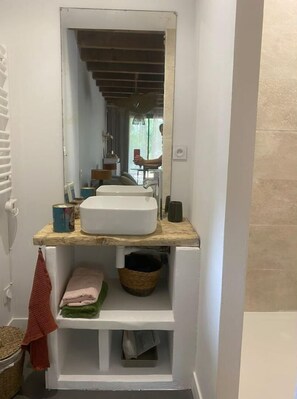 Une salle de bain avec un lavabo blanc, un miroir au-dessus et une étagère en bois en dessous. Il y a un porte-serviettes à gauche et un panier à droite.