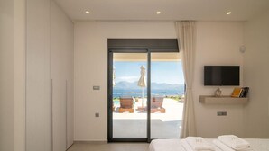 A hotel room with a bed, a TV mounted on the wall, and a view of a pool and mountains through a sliding door. - Horizon View Villa with Endless View (Agios Nikolaos)