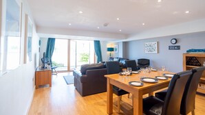 A modern living room with a dining area, wooden flooring, and a large window with curtains. - Fistral Beach - Apartment 3 (Newquay)
