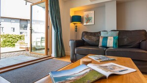 A modern living room with a leather sofa, a wooden coffee table with magazines, a floor lamp, and a sliding glass door leading to an outdoor area with a garden. - Fistral Beach - Apartment 3 (Newquay)
