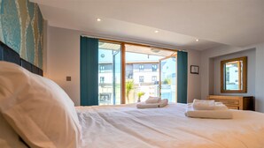 A hotel room with a large bed, a wooden nightstand, a mirror, and a view of the outside buildings through a large window. - Fistral Beach - Apartment 3 (Newquay)