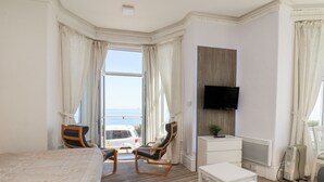A hotel room with a bed, two chairs, a small table, a TV, and a view of the sea.
