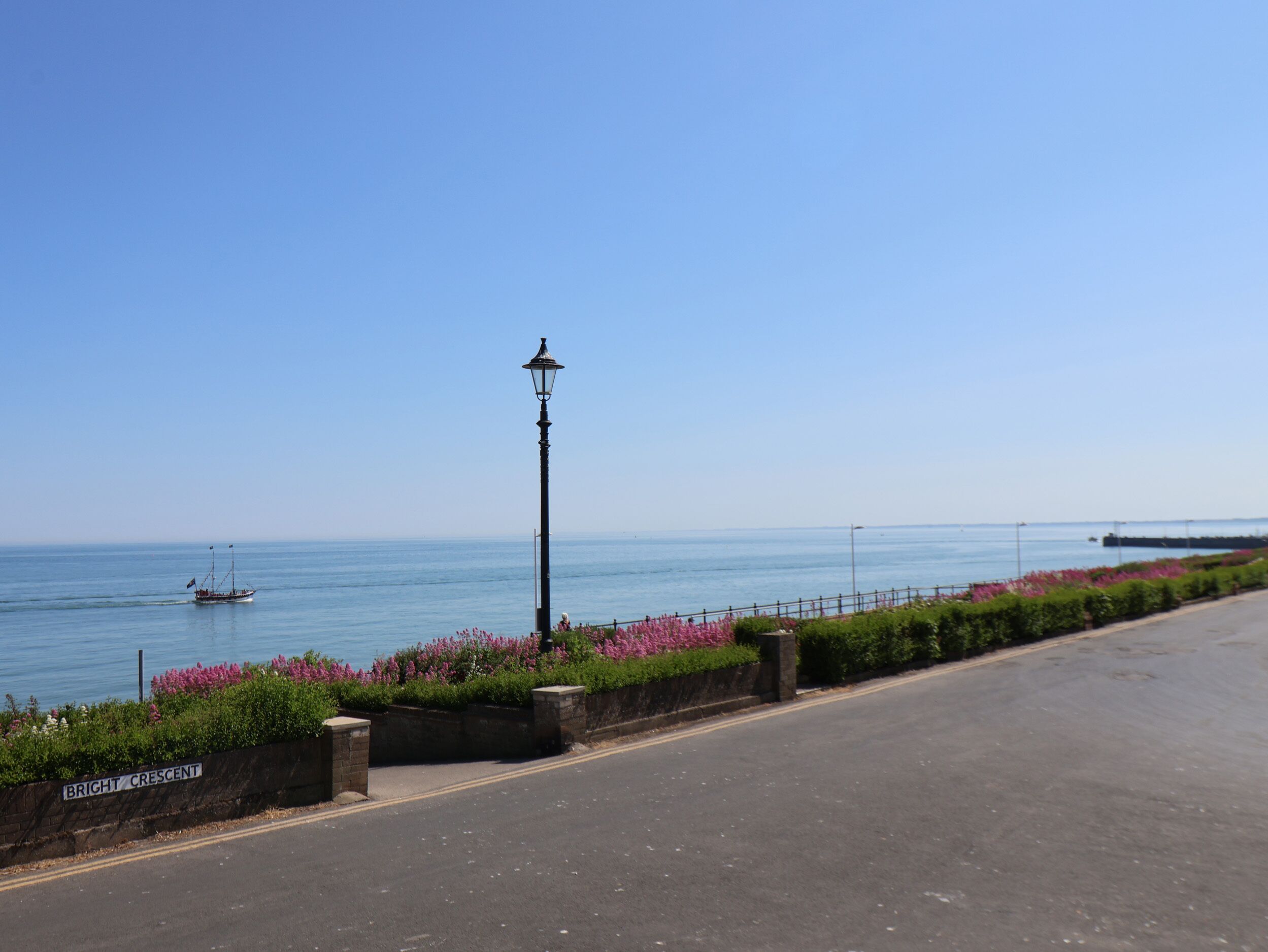 A coastal road with a street lamp, a sign reading "Bright Crescent," and a view of the sea with a boat in the distance.