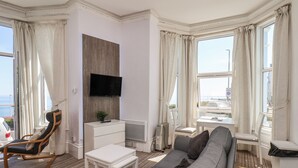 A modern living room with a grey sofa, a white side table, a wooden chair, a TV mounted on a wooden panel, and large windows with curtains.