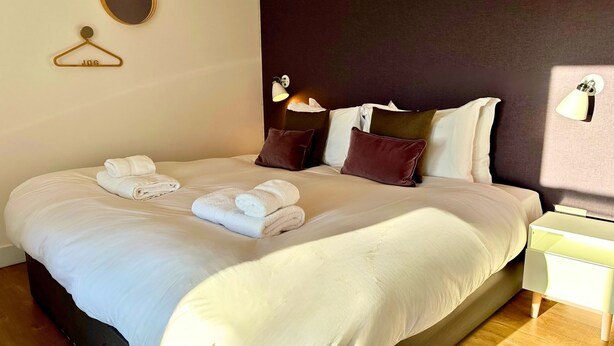 Een net opgemaakt bed met twee handdoeken, een donker hoofdeinde en bedlampjes.