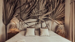 Una cama con cabecera hecha de grandes hojas de palma secas, dos almohadas y una mesita de noche con una lámpara.