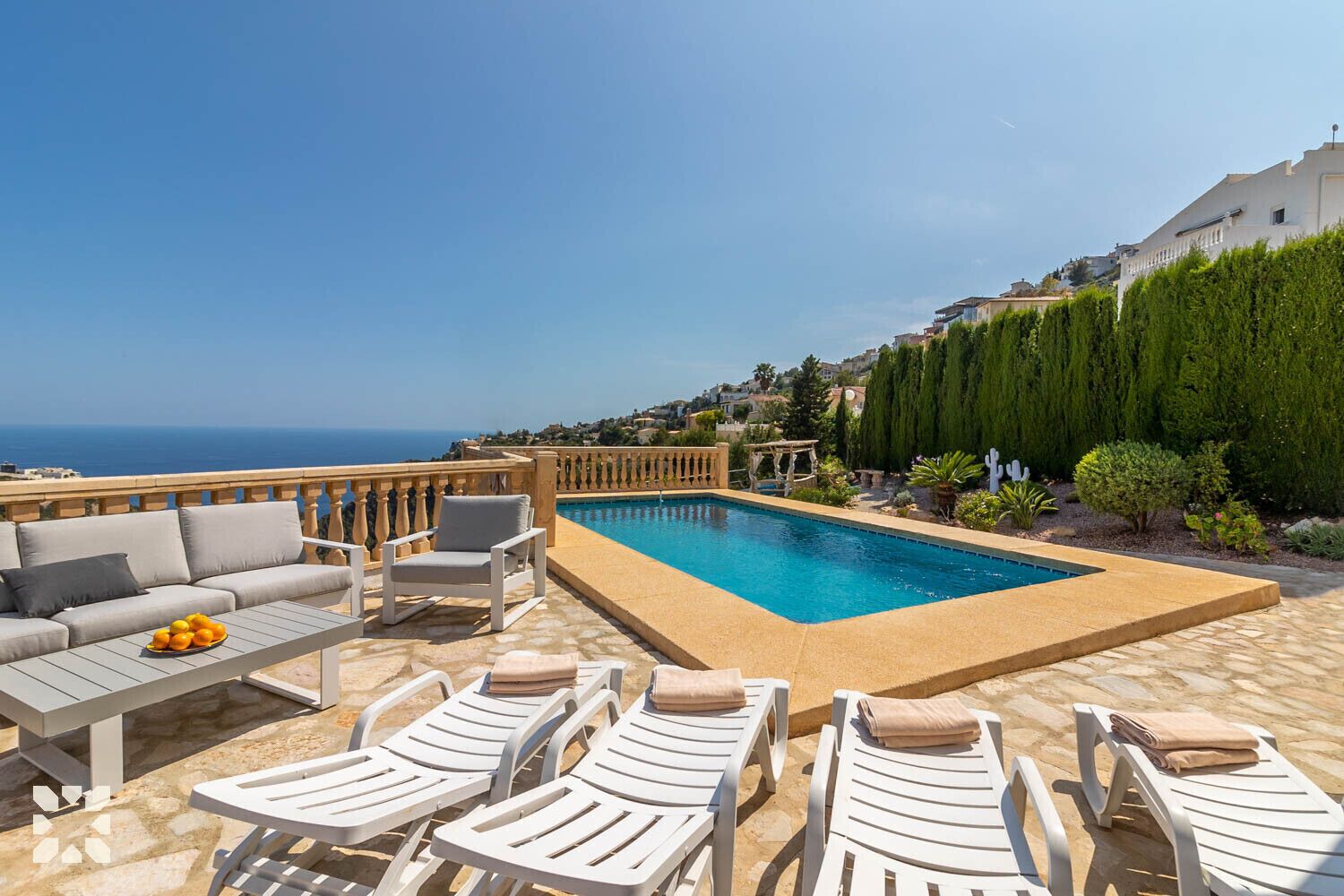 A pool area with lounge chairs and a sofa, surrounded by greenery and overlooking a coastal view.