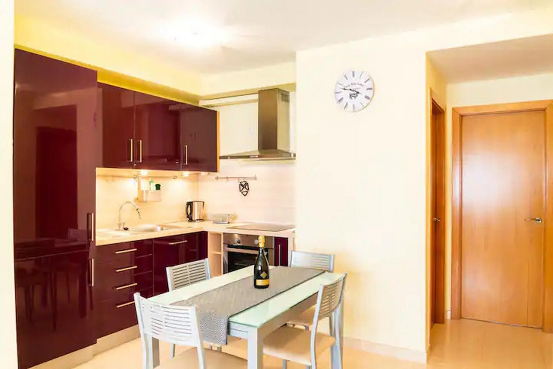 A compact kitchen with dark cabinets, a dining table with chairs, a clock on the wall, and a wooden door.