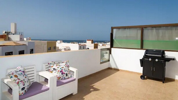 Une terrasse sur le toit avec un canapé blanc, des coussins colorés, un barbecue et une vue sur la mer.