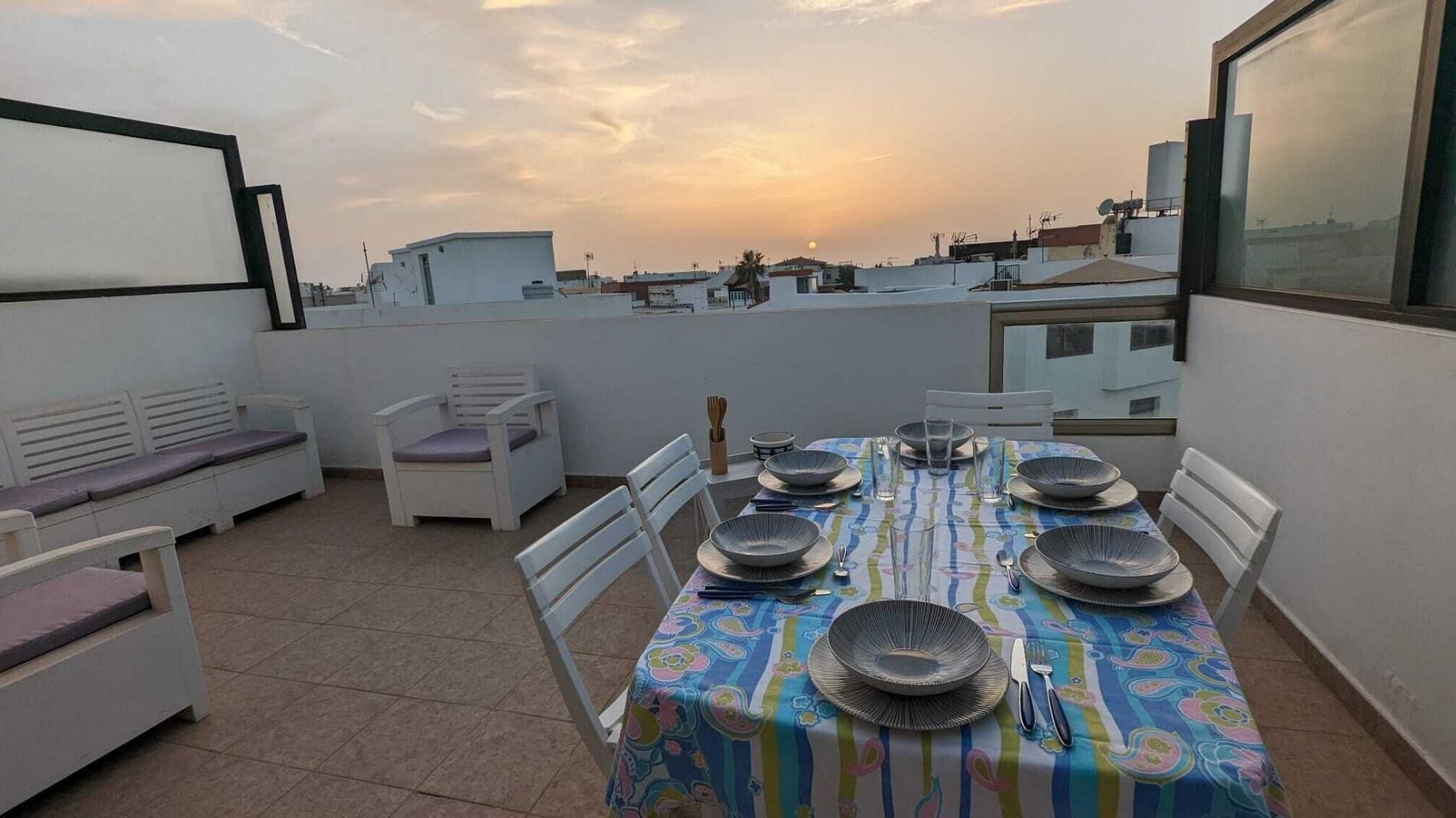 A rooftop terrace with a dining table set for six, surrounded by white chairs, and a view of the cityscape at sunset.