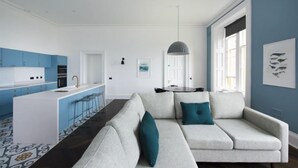 A modern living room with a sofa and a dining table, a kitchen with blue cabinets, and a large window with artwork. - Portrona (Isle of Lewis)