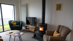 Een gezellige woonkamer met een open haard, een groene fauteuil, een bruine fauteuil, een televisie en een houten bijzettafeltje.