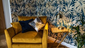 Een mosterdgele bank met een donkerblauwe kussen, een klein houten bijzettafeltje met een lamp, en een pot met een plant.