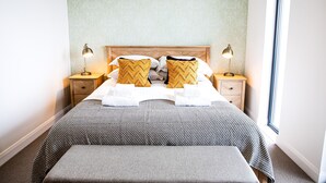 Een opgemaakt bed met een beddekleed met dessin, twee kussens met een ruitpatroon, en een passend bankje. Er staan twee bedlampen op houten nachtkastjes.