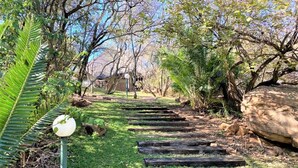 A pathway with wooden planks leading through a garden with trees and a large rock on the right.