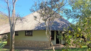 A building with a stone foundation and a thatched roof, surrounded by a grassy area and trees.